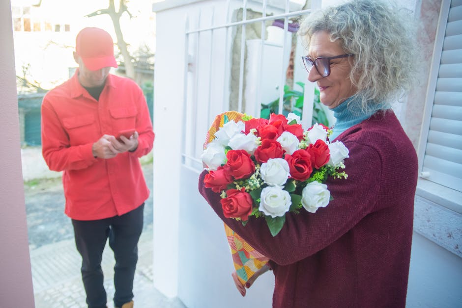 A woman with curly gray hair and glasses holding a bouquet of fresh red and white roses, wrapped in colorful paper, while standing on a porch. A delivery person in a red jacket and cap is in the backg