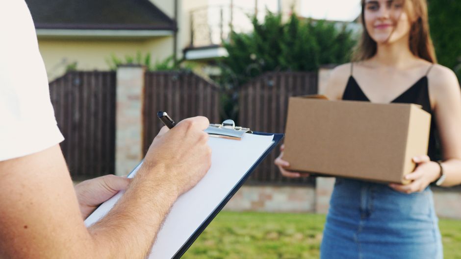 A woman standing outdoors in a garden holding a plain cardboard box, possibly containing floral gifts, while a person off-camera writes on a clipboard with a pen. The scene is well-lit with natural da