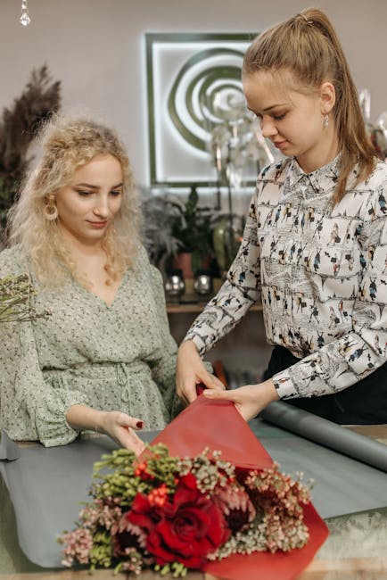 A floral arrangement being crafted by a professional florist in an indoor setting. The bouquet features vibrant red roses complemented by pink and white baby's breath, all arranged with lush green fol
