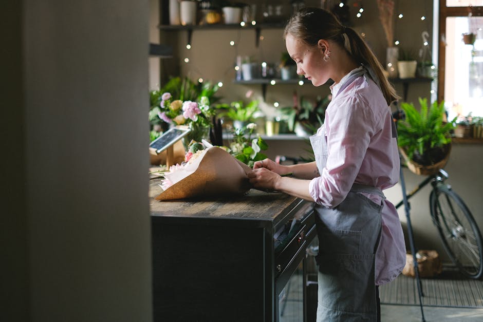 A young female florist with light brown hair tied back, wearing a light pink shirt and a grey apron, is carefully arranging a bouquet of fresh flowers on a wooden workbench inside a modern floral shop