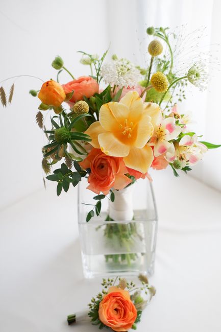 A vibrant floral arrangement in a clear glass vase featuring large yellow lisianthus and soft orange roses, complemented by pink and white alstroemeria, small round green amaranthus, and delicate whit