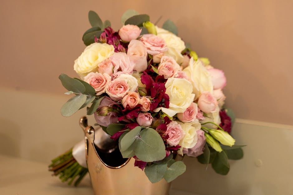 A bouquet arrangement featuring a harmonious blend of soft pink and creamy white roses, complemented by deep burgundy blooms and lush green eucalyptus leaves. The flowers are tightly clustered, with t